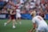 A sporting event with athletes on a grass field, one in a white jersey with the number 11 in the foreground, holding a lacrosse stick. Players in the background wear maroon uniforms. Spectators fill the stands creating a lively atmosphere.