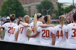 A group of female athletes wearing white sports jerseys with red numbers stands closely together, some with arms around each other. They appear to be preparing for or participating in a sporting event on a field, with a blurred background of spectators and trees.