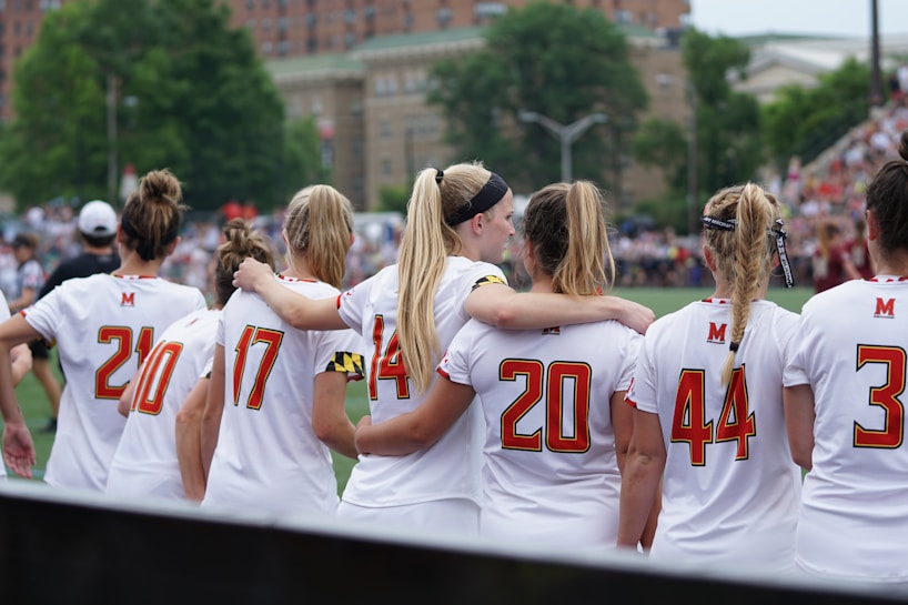 A group of female athletes wearing white sports jerseys with red numbers stands closely together, some with arms around each other. They appear to be preparing for or participating in a sporting event on a field, with a blurred background of spectators and trees.