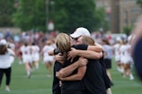 A group of three people embrace in a heartfelt hug on a sports field with a blurred background of players and officials in athletic attire. The individuals appear to be wearing casual clothing, and the atmosphere suggests celebration or camaraderie.