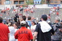 A lively gathering in an outdoor sports venue with a group of people, some wearing matching shirts labeled 'National Champions.' Several individuals are holding cameras and microphones, indicating a media presence. The background shows blurred spectators sitting on bleachers.