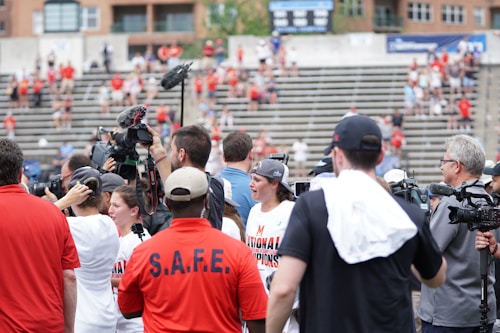 A lively gathering in an outdoor sports venue with a group of people, some wearing matching shirts labeled 'National Champions.' Several individuals are holding cameras and microphones, indicating a media presence. The background shows blurred spectators sitting on bleachers.