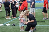 A young child wearing a sports t-shirt stands next to an adult who is crouching down. The scene takes place on a sports field with several people in casual sportswear milling around in the background.