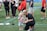 A young child wearing a sports t-shirt stands next to an adult who is crouching down. The scene takes place on a sports field with several people in casual sportswear milling around in the background.