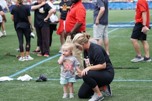 A young child wearing a sports t-shirt stands next to an adult who is crouching down. The scene takes place on a sports field with several people in casual sportswear milling around in the background.