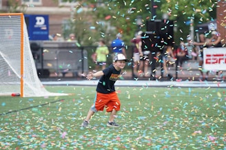 A child wearing a black T-shirt, orange shorts, and a cap is playfully running on a sports field amidst a shower of colorful confetti. A lacrosse goal is visible in the background alongside blurred figures and a banner.