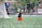 A child wearing a black T-shirt, orange shorts, and a cap is playfully running on a sports field amidst a shower of colorful confetti. A lacrosse goal is visible in the background alongside blurred figures and a banner.