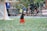 A child wearing a black T-shirt, orange shorts, and a cap is playfully running on a sports field amidst a shower of colorful confetti. A lacrosse goal is visible in the background alongside blurred figures and a banner.