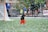 A child wearing a black T-shirt, orange shorts, and a cap is playfully running on a sports field amidst a shower of colorful confetti. A lacrosse goal is visible in the background alongside blurred figures and a banner.