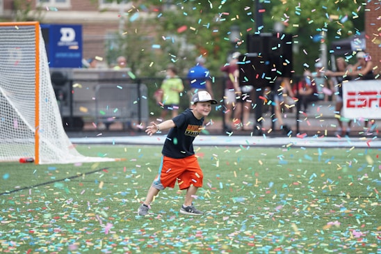 A child wearing a black T-shirt, orange shorts, and a cap is playfully running on a sports field amidst a shower of colorful confetti. A lacrosse goal is visible in the background alongside blurred figures and a banner.