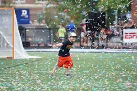 A child wearing a black T-shirt, orange shorts, and a cap is playfully running on a sports field amidst a shower of colorful confetti. A lacrosse goal is visible in the background alongside blurred figures and a banner.