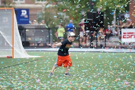 A child wearing a black T-shirt, orange shorts, and a cap is playfully running on a sports field amidst a shower of colorful confetti. A lacrosse goal is visible in the background alongside blurred figures and a banner.