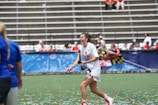 A woman in sports attire is joyfully running on a field while holding a trophy. Confetti is scattered on the ground, and there are people in the background seated in bleachers, some of whom are cheering. The woman is wearing a white shirt with 'National Champions' printed on it.