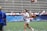 A woman in sports attire is joyfully running on a field while holding a trophy. Confetti is scattered on the ground, and there are people in the background seated in bleachers, some of whom are cheering. The woman is wearing a white shirt with 'National Champions' printed on it.