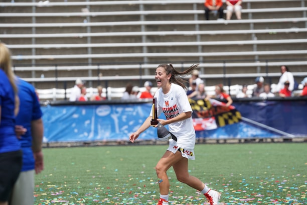 A woman in sports attire is joyfully running on a field while holding a trophy. Confetti is scattered on the ground, and there are people in the background seated in bleachers, some of whom are cheering. The woman is wearing a white shirt with 'National Champions' printed on it.