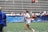 A woman in sports attire is joyfully running on a field while holding a trophy. Confetti is scattered on the ground, and there are people in the background seated in bleachers, some of whom are cheering. The woman is wearing a white shirt with 'National Champions' printed on it.