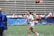 A woman in sports attire is joyfully running on a field while holding a trophy. Confetti is scattered on the ground, and there are people in the background seated in bleachers, some of whom are cheering. The woman is wearing a white shirt with 'National Champions' printed on it.