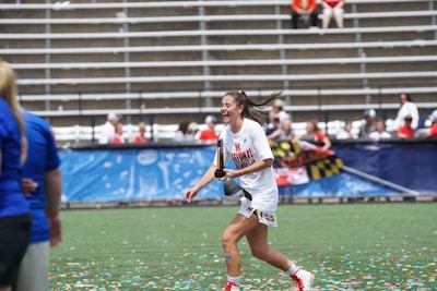 A woman in sports attire is joyfully running on a field while holding a trophy. Confetti is scattered on the ground, and there are people in the background seated in bleachers, some of whom are cheering. The woman is wearing a white shirt with 'National Champions' printed on it.