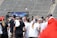 A group of athletes in white shirts with black and red accents are gathered together, engaged in conversation and smiling. They appear to be on a sports field with empty bleachers in the background. The setting is outdoors, and it seems sunny. Some individuals are wearing caps or headbands.