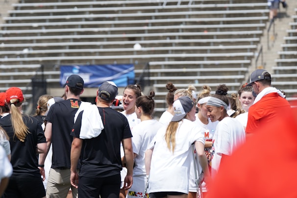 A group of athletes in white shirts with black and red accents are gathered together, engaged in conversation and smiling. They appear to be on a sports field with empty bleachers in the background. The setting is outdoors, and it seems sunny. Some individuals are wearing caps or headbands.