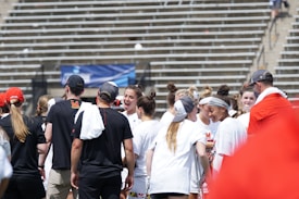 A group of athletes in white shirts with black and red accents are gathered together, engaged in conversation and smiling. They appear to be on a sports field with empty bleachers in the background. The setting is outdoors, and it seems sunny. Some individuals are wearing caps or headbands.