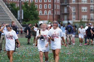 A group of young women, wearing 'National Champions' t-shirts, walk across a field covered with colorful confetti. They appear to be celebrating a victory. The background shows a few people mingling and large brick buildings.
