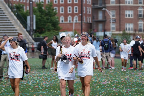 A group of young women, wearing 'National Champions' t-shirts, walk across a field covered with colorful confetti. They appear to be celebrating a victory. The background shows a few people mingling and large brick buildings.