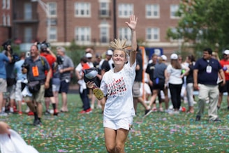 A jubilant person wearing sports attire is smiling and waving while holding a cap. They are standing on a field strewn with confetti, surrounded by a crowd of people who appear to be celebrating. The background shows buildings and greenery.
