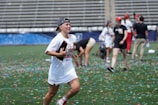 A person wearing a championship T-shirt and a cap runs joyfully on a field covered with colorful confetti. In the background, people are celebrating, with some bending down to pick up items. The scene takes place in a stadium with empty bleachers.