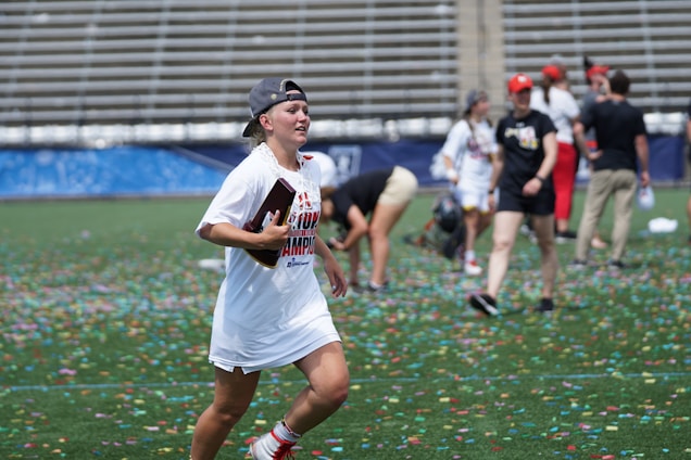 A person wearing a championship T-shirt and a cap runs joyfully on a field covered with colorful confetti. In the background, people are celebrating, with some bending down to pick up items. The scene takes place in a stadium with empty bleachers.
