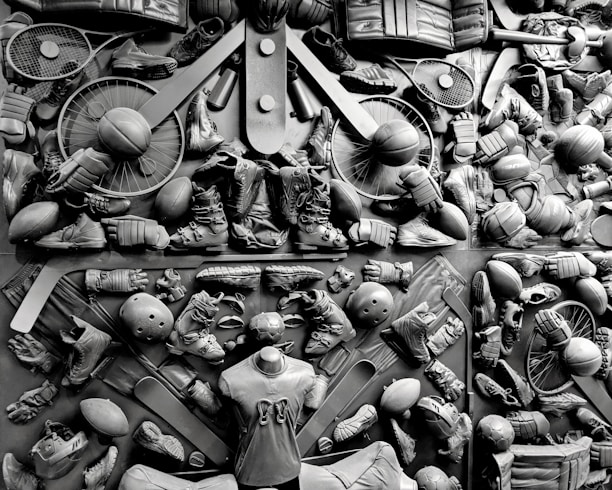 A complex arrangement of various sports equipment and attire, including tennis rackets, soccer balls, basketballs, helmets, shoes, gloves, and a mannequin torso wearing a T-shirt. The items are layered and artistically displayed in a monochrome color scheme, creating an intricate texture and visual pattern.