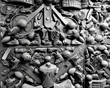 A complex arrangement of various sports equipment and attire, including tennis rackets, soccer balls, basketballs, helmets, shoes, gloves, and a mannequin torso wearing a T-shirt. The items are layered and artistically displayed in a monochrome color scheme, creating an intricate texture and visual pattern.