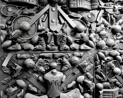 A complex arrangement of various sports equipment and attire, including tennis rackets, soccer balls, basketballs, helmets, shoes, gloves, and a mannequin torso wearing a T-shirt. The items are layered and artistically displayed in a monochrome color scheme, creating an intricate texture and visual pattern.