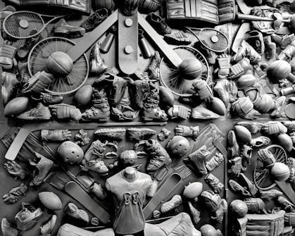 A complex arrangement of various sports equipment and attire, including tennis rackets, soccer balls, basketballs, helmets, shoes, gloves, and a mannequin torso wearing a T-shirt. The items are layered and artistically displayed in a monochrome color scheme, creating an intricate texture and visual pattern.