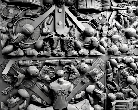 A complex arrangement of various sports equipment and attire, including tennis rackets, soccer balls, basketballs, helmets, shoes, gloves, and a mannequin torso wearing a T-shirt. The items are layered and artistically displayed in a monochrome color scheme, creating an intricate texture and visual pattern.