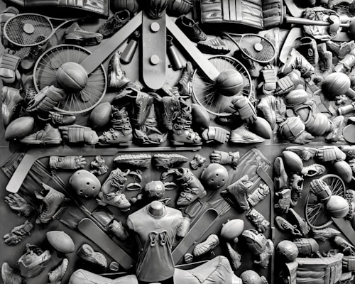 A complex arrangement of various sports equipment and attire, including tennis rackets, soccer balls, basketballs, helmets, shoes, gloves, and a mannequin torso wearing a T-shirt. The items are layered and artistically displayed in a monochrome color scheme, creating an intricate texture and visual pattern.
