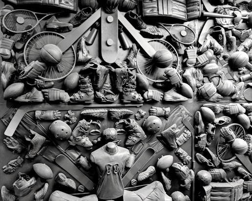 A complex arrangement of various sports equipment and attire, including tennis rackets, soccer balls, basketballs, helmets, shoes, gloves, and a mannequin torso wearing a T-shirt. The items are layered and artistically displayed in a monochrome color scheme, creating an intricate texture and visual pattern.