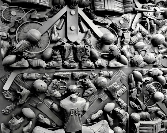 A complex arrangement of various sports equipment and attire, including tennis rackets, soccer balls, basketballs, helmets, shoes, gloves, and a mannequin torso wearing a T-shirt. The items are layered and artistically displayed in a monochrome color scheme, creating an intricate texture and visual pattern.