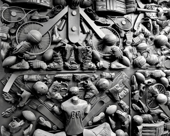 A complex arrangement of various sports equipment and attire, including tennis rackets, soccer balls, basketballs, helmets, shoes, gloves, and a mannequin torso wearing a T-shirt. The items are layered and artistically displayed in a monochrome color scheme, creating an intricate texture and visual pattern.