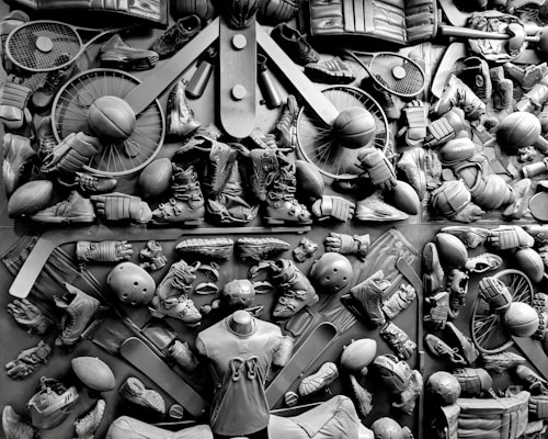 A complex arrangement of various sports equipment and attire, including tennis rackets, soccer balls, basketballs, helmets, shoes, gloves, and a mannequin torso wearing a T-shirt. The items are layered and artistically displayed in a monochrome color scheme, creating an intricate texture and visual pattern.
