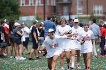 A group of athletes in white apparel with 'National Champions' printed on their shirts celebrate on a field covered in confetti. They hold trophies and wear hats. Spectators and photographers can be seen in the background, capturing the festive atmosphere.