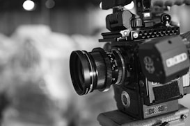 A close-up of a professional video camera with a detailed lens and various controls and attachments. The image is in black and white, highlighting the intricate design and mechanics of the equipment.