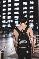 A person stands with their back to the viewer, wearing a beige shirt and a black backpack. The backpack features the word 'Brooklyn' and an NBA logo. The setting is urban and appears to be at night, with a tall building in the background illuminated by numerous lit windows.