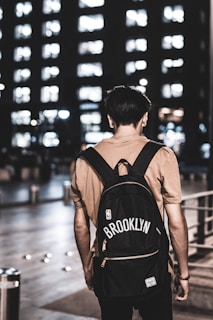 A person stands with their back to the viewer, wearing a beige shirt and a black backpack. The backpack features the word 'Brooklyn' and an NBA logo. The setting is urban and appears to be at night, with a tall building in the background illuminated by numerous lit windows.