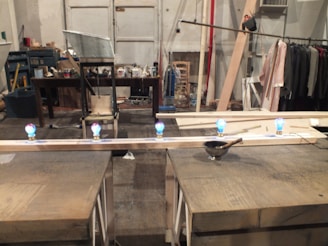 A workspace with wooden beams and small colorful lights on top of them. The area appears cluttered with tools and materials scattered around. In the background, there is a table filled with various items and some clothing hanging on racks.