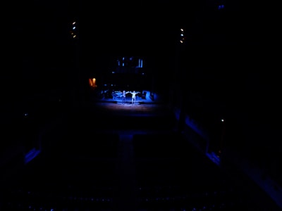 A dark theater setting with a brightly lit stage. Three performers stand in the center, illuminated by spotlights. The rest of the auditorium appears dimly lit, highlighting the contrast between the stage and the audience area.