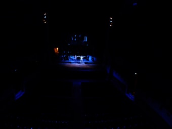 A dark theater setting with a brightly lit stage. Three performers stand in the center, illuminated by spotlights. The rest of the auditorium appears dimly lit, highlighting the contrast between the stage and the audience area.