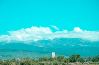 A scenic landscape features a range of distant mountains beneath a clear, bright blue sky filled with fluffy white clouds. In the foreground, a sign indicating 'Terrain à Vendre' is visible, surrounded by grass and scrubland with some sparse trees. A fence runs along the landscape, adding a rustic touch to the serene environment.