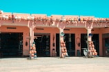 A market stall displays a variety of pottery items, including pots, vases, and decorative items arranged neatly on stands and shelves. The building features a pink facade with arches, and the atmosphere suggests it is a sunny day with a clear blue sky. Several pottery items are also lined up on the roof. A scooter is parked in front of the stall, and the ground appears dusty.