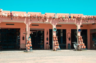 A market stall displays a variety of pottery items, including pots, vases, and decorative items arranged neatly on stands and shelves. The building features a pink facade with arches, and the atmosphere suggests it is a sunny day with a clear blue sky. Several pottery items are also lined up on the roof. A scooter is parked in front of the stall, and the ground appears dusty.